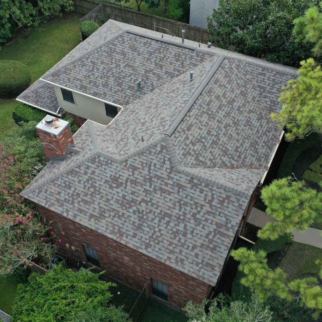 Aerial view of a house with an asphalt shingle roof, showcasing a brick chimney and multiple roof sections. The house is surrounded by green trees, shrubs, and a fenced yard. Expertly installed by a roofing company in Memorial City, the shingles blend seamlessly in varying shades of gray with the brick exterior.