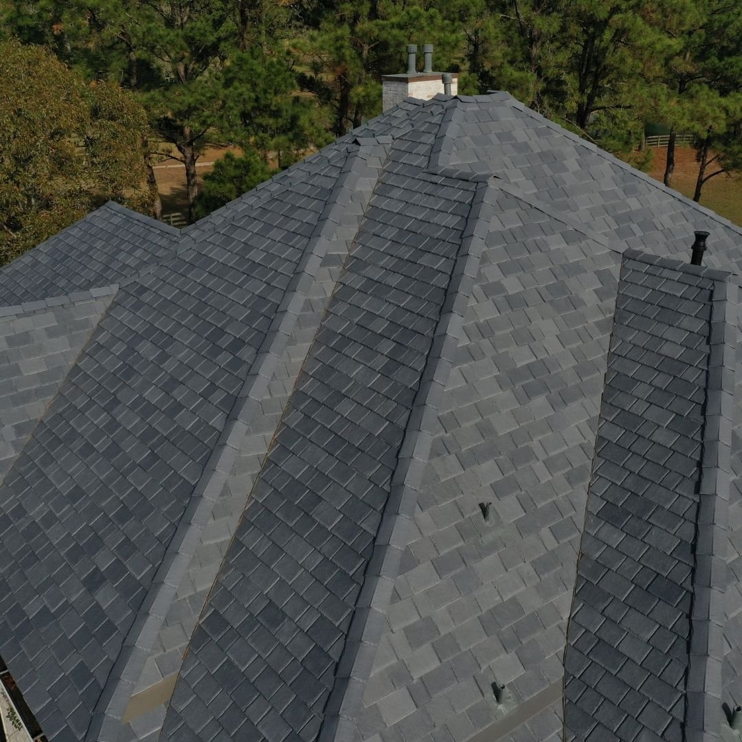 Aerial view of a large roof with dark gray, textured shingles installed by a roofing company in Memorial City. Multiple ridges and valleys can be seen, as well as a chimney near the center. Surrounding the house are tall trees.