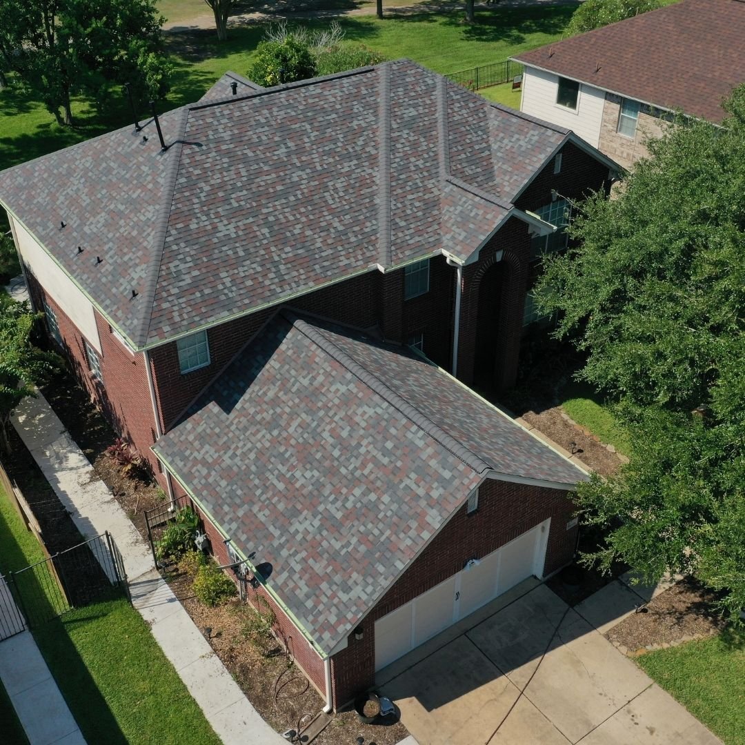 Aerial view of a two-story brick house with a multi-faceted gray shingle roof, expertly installed by a roofing company in Memorial City, and an attached two-car garage. The surrounding area includes a driveway, green lawns, and adjacent homes. Trees and a fence line the property, providing a well-maintained appearance.