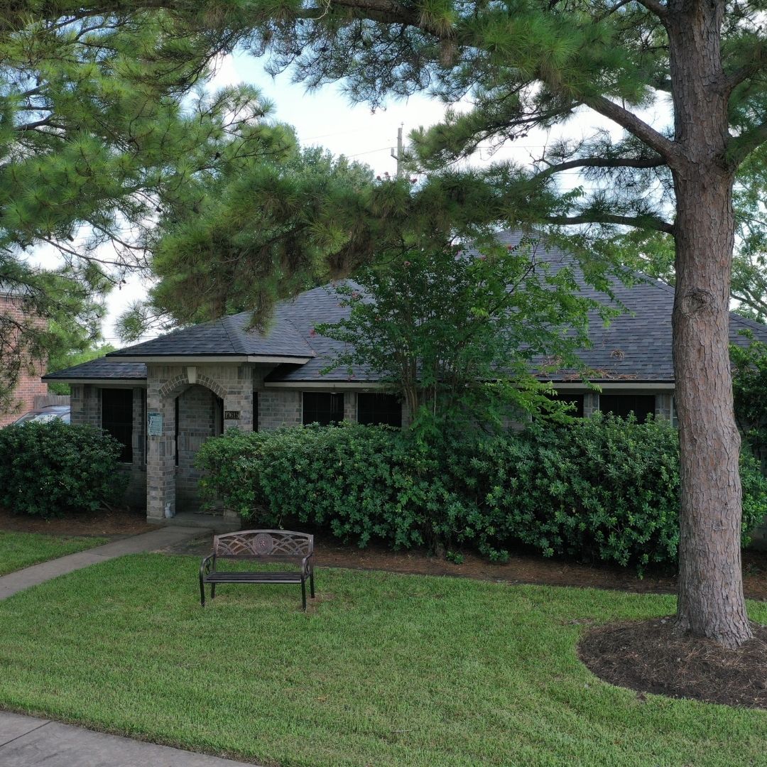 A single-story house with gray brickwork is partially obscured by lush greenery. A large tree shades the front lawn, which also features a wooden bench near a paved walkway leading to the entrance. The roof, expertly installed by a roofing company in Memorial City, is dark gray.