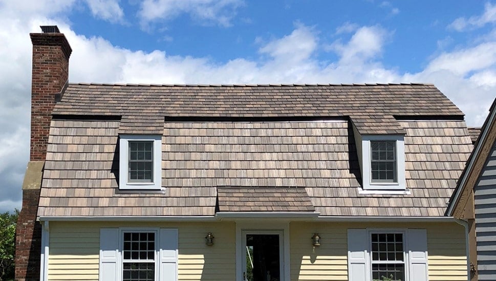 A two-story house with light yellow siding, white trim, and a wood-shingled Gambrel roof. The roof has two dormer windows with white frames, and there is a central entrance with a small overhanging roof. A cloudy blue sky is in the background.
