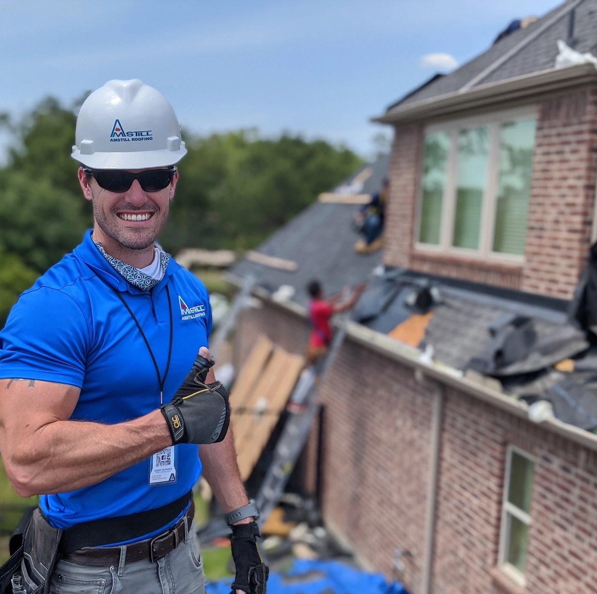 A man wearing a white hard hat with the logo "ACC" and a blue shirt smiles and gives a thumbs-up gesture at a construction site. Workers are seen in the background working on the roof of a brick house. The weather is clear and sunny.