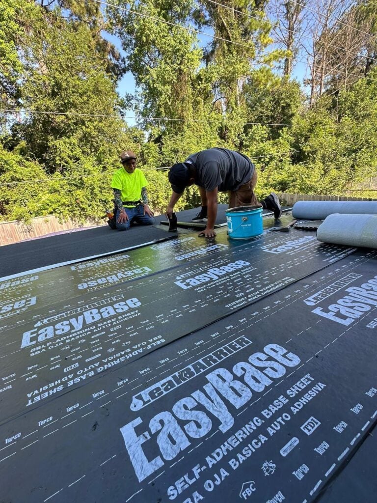 Two workers on a roof installing a self-adhering base sheet. One is kneeling and applying the sheet, while the other sits nearby. Trees are in the background. The sheet is labeled "EasyBase.