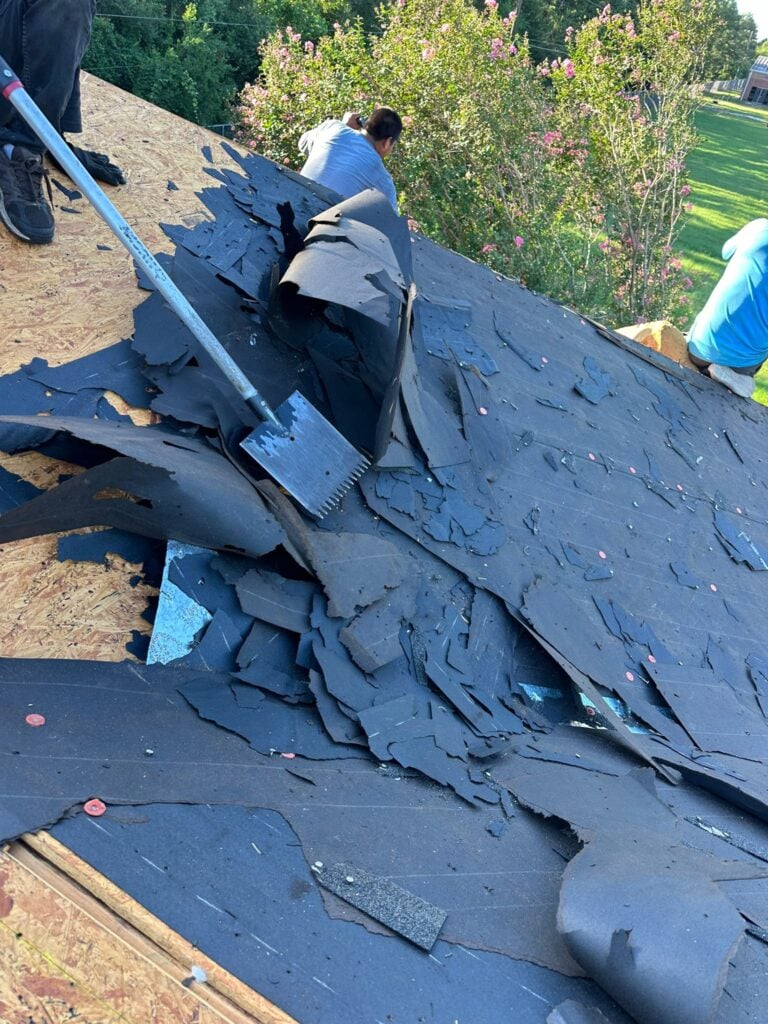 Workers are removing old, damaged roofing materials from a wooden roof. One worker uses a long scraper tool, while another is visible in the background. Nearby trees and a grassy area can be seen. The sky is clear, suggesting a sunny day.
