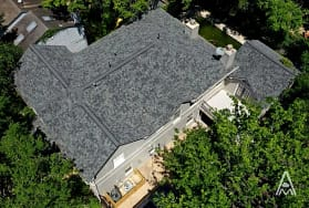 Aerial view of a large house with a complex, dark gray shingled roof surrounded by lush green trees. A wooden deck is visible on one side. The property is nestled in a residential area with neighboring homes partially visible.