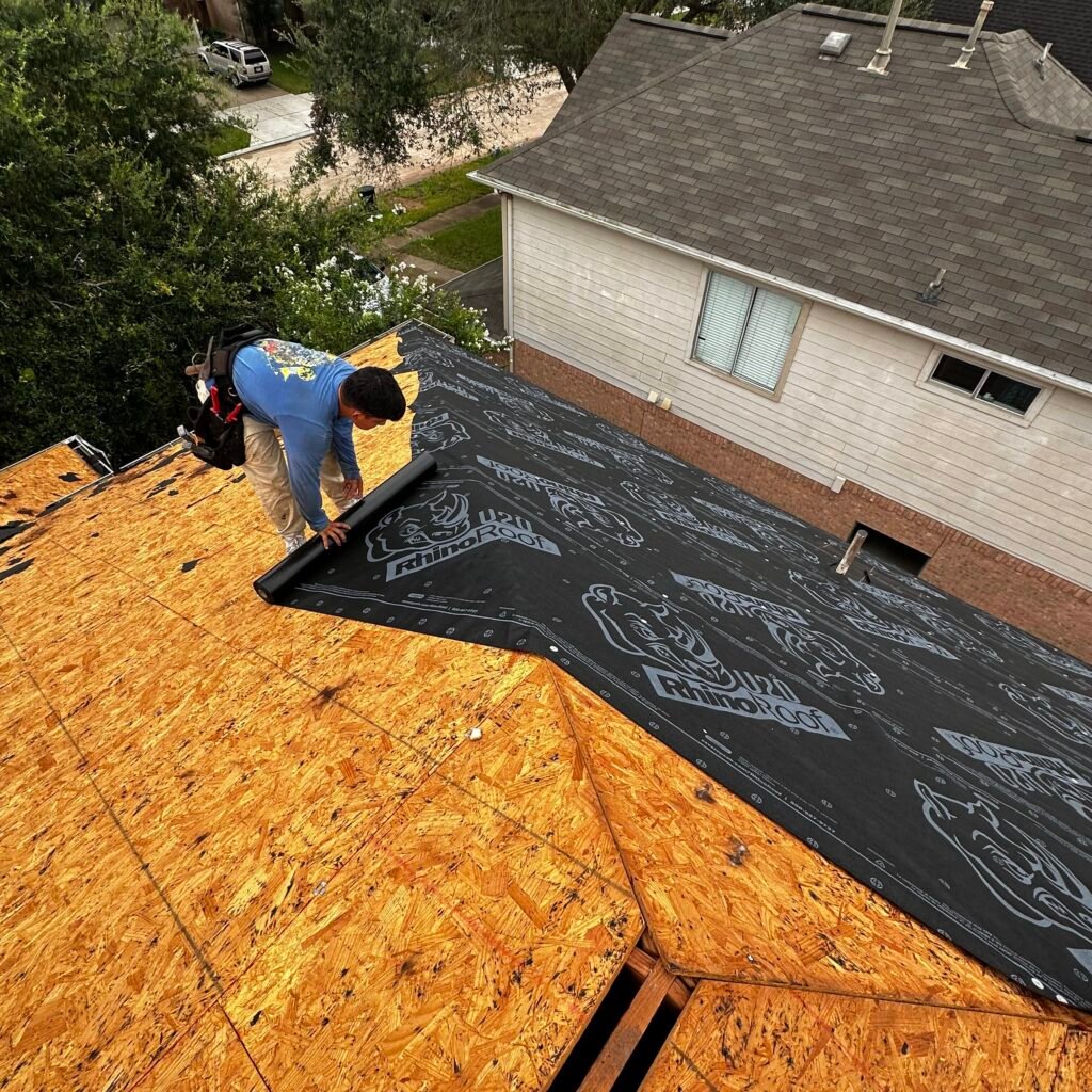 A person in a blue shirt is diligently installing roofing underlayment on a house roof made of wooden panels in Houston. The scene captures the partially covered roof with nearby trees as a backdrop, highlighting the meticulous Houston roofing craftsmanship involved.
