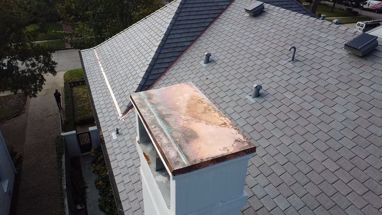 A rooftop scene showing a gray tiled roof with a shiny copper chimney cap. The roof has several small vents and a triangular design. A person is visible walking on the sidewalk near the house. Trees and a street are seen in the background.
