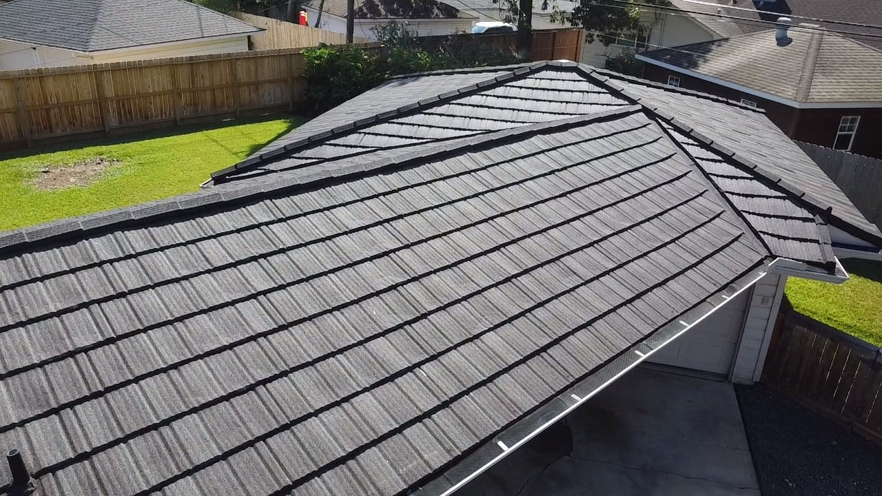 Aerial view of a residential roof with dark shingles and a textured pattern. The roof is in a neighborhood with green lawns and wooden fences, and covers a garage partially visible below.