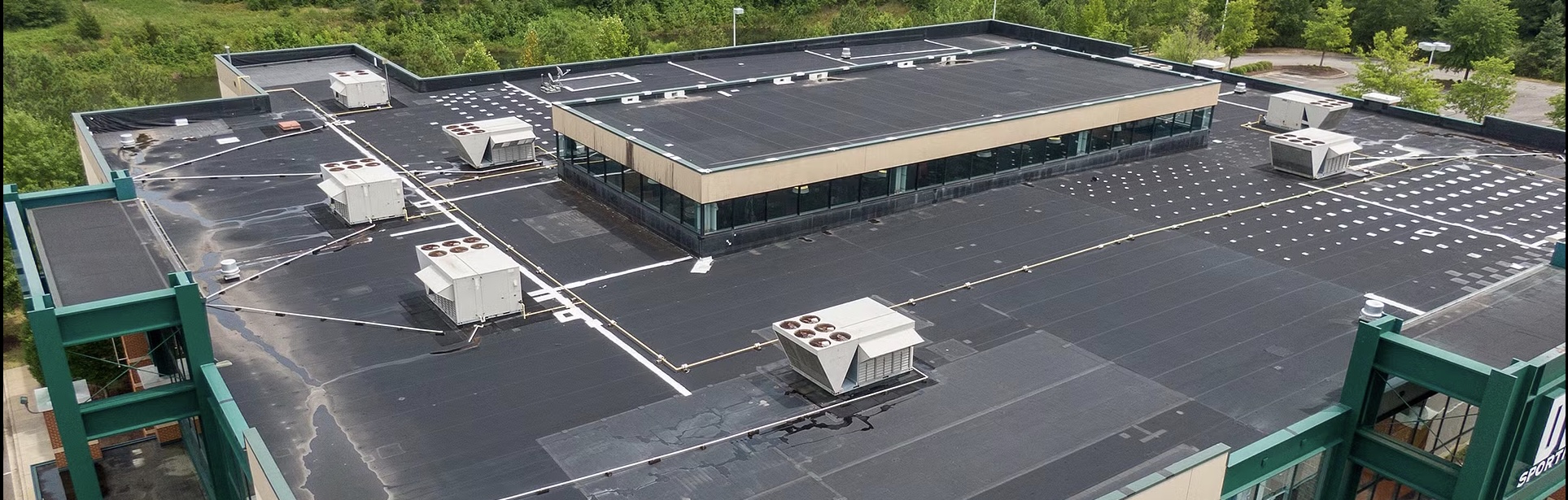 Aerial view of a flat commercial building roof with multiple HVAC units, surrounded by trees and greenery, showing roofing seams and patches across the black surface.