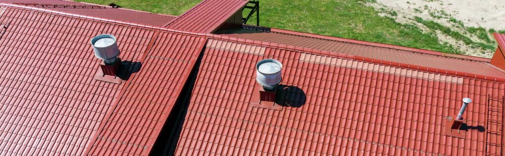 Aerial view of a commercial red metal roof with two silver rooftop vents, green grass visible along the roof’s edge.